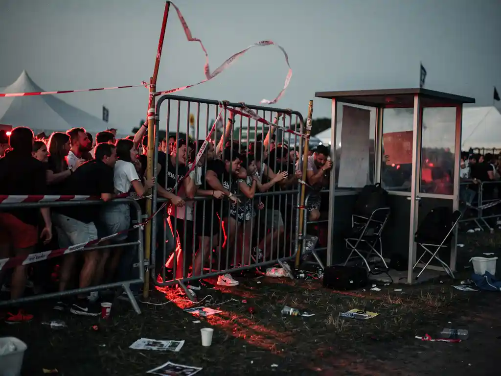 Menigte die bij schemering tegen een onbemand festivalhek dringt, met losgeraakte afzetlinten en verlaten beveiligingspost.
