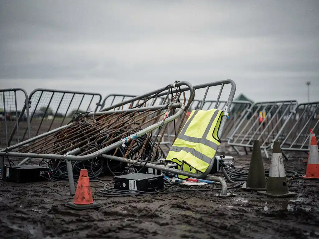 Ingestort festivalpodiumbord met verspreid crowd-control hekwerk en een vergeten veiligheidsvest op modderig buitenterrein.